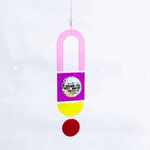 A hanging decorative ornament shaped like a stylized U with a pink top, a purple middle section featuring a mirrored disco ball, a yellow semicircle, and a red semicircle at the bottom, shown against a soft white background.