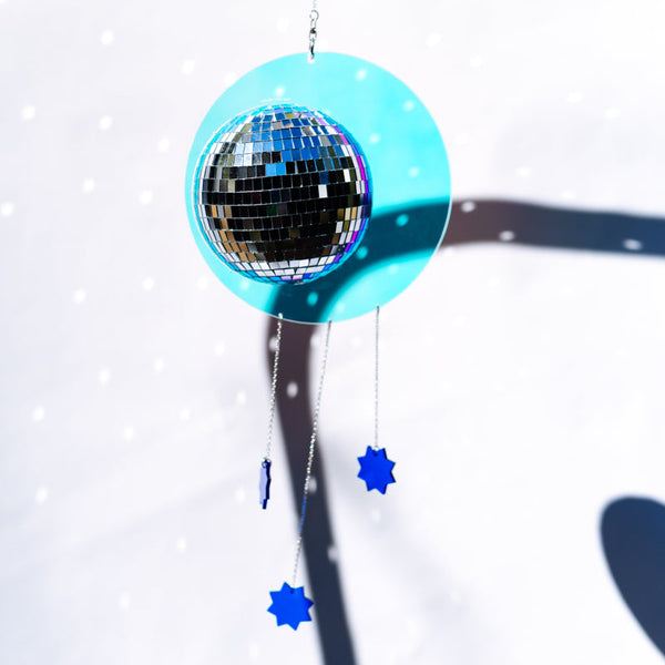A hanging disco ball ornament with a bright turquoise circular frame and mirrored center sphere, suspended by a thin cord against a white background with soft shadows and small light specks.