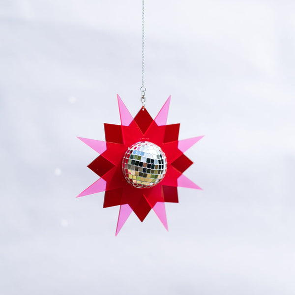A hanging star-shaped disco ball ornament with layered red and pink paper petals surrounding a reflective silver mirrored ball, suspended against a soft white background.
