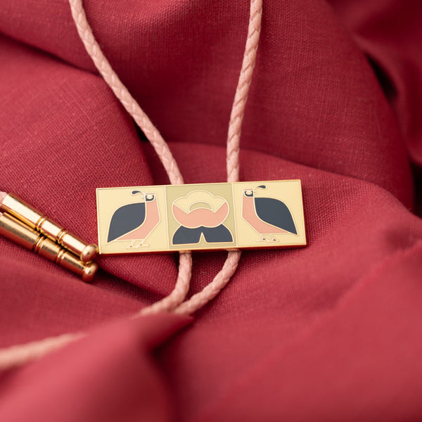 a hard enamel bolo tie featuring an illustration of two California quails and a flower, with pink vegan cord and gold metal tips on a maroon fabric background