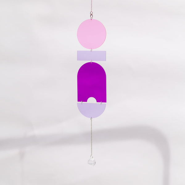 A minimalist acrylic wind spinner ornament featuring geometric shapes in pink, purple, and lavender, suspended by a thin chain with a small clear crystal bead at the bottom.